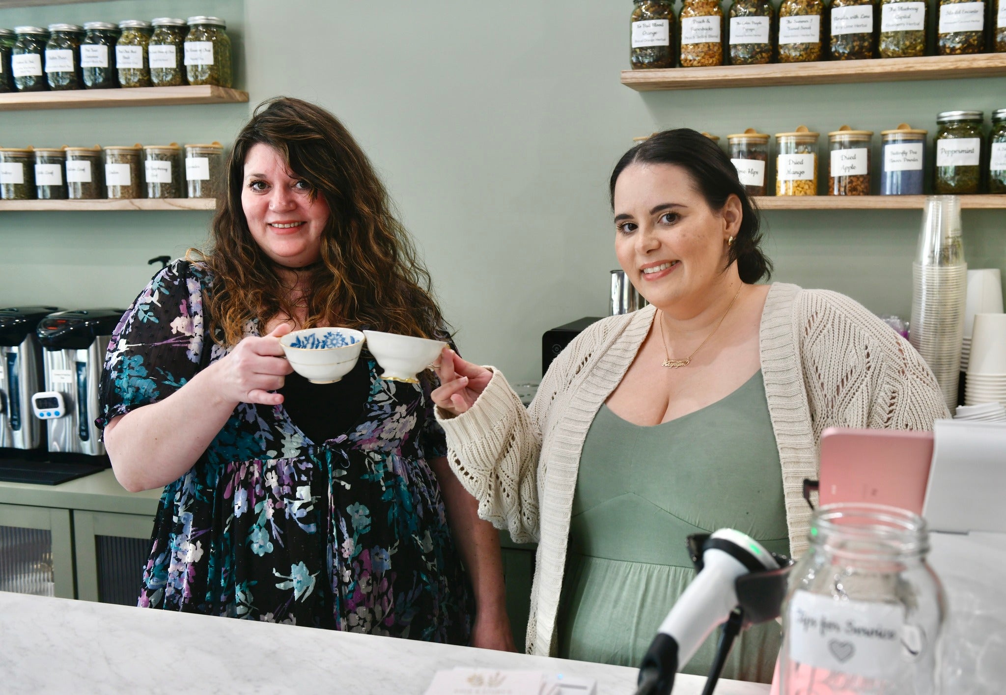 Khrystyne and Cassandra raising teacups behind the tea bar at Sage & Story's Teahouse